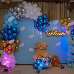 A classic "Welcome Home Baby" decoration designed for an indoor space. This setup includes a large teddy bear, a custom neon "Welcome" sign, and a beautiful balloon arch in shades of blue, silver, and gold against a sky-themed backdrop.
