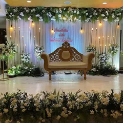 An elegant reception stage in white and green. The backdrop features white drapes with hanging industrial bulbs, framed by beautiful white floral arrangements.