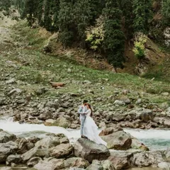 A wide shot showing the couple standing on a boulder in the middle of a river in Kashmir. This image captures the scale of the landscape and the adventurous spirit of the couple.