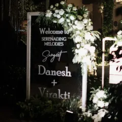 A sleek and modern welcome sign for the "Serenading Melodies" Sangeet. The black acrylic with white lettering, adorned with fresh white flowers, set a sophisticated tone right from the entrance.