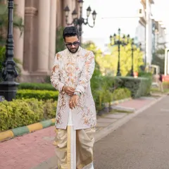 This groom is pictured in a custom off-white sherwani with elaborate gold and white embroidery. The outfit is paired with a silk dhoti, creating a classic and sophisticated look suitable for a wedding ceremony.