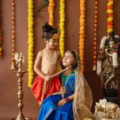 A sweet interaction between siblings dressed as Krishna and Radha. The setup includes a traditional swing, marigold garlands, and brass decor to create an authentic festive atmosphere.
