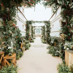 A rustic and lush walkway for a nature-inspired wedding. The path is lined with an abundance of ferns, tropical leaves, and wooden crates, creating an enchanted forest feel.