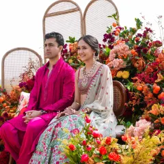 A happy couple at their sun-kissed Haldi in Udaipur. They are seated amidst a stunning explosion of citrus-hued florals, including oranges, pinks, and reds, creating a vibrant and joyful backdrop.