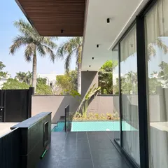 This view from the covered veranda at Gulmohar Farmhouse shows how the pool is integrated directly with the living space. The wooden ceiling and dark floor tiles create a warm, resort-like feel, with views of palm trees framing the scene.