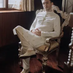 A relaxed portrait of the groom seated in an ornate, gilded chair, looking confidently at the camera in his elegant white sherwani.