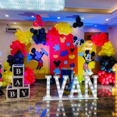 A vibrant and grand Mickey Mouse themed first birthday. This large-scale setup includes multiple character backdrops, colorful balloon arrangements, light-up letters for the name 'IVAN', and a marquee number '1'.