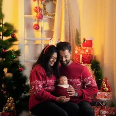 A cozy family portrait with parents and their newborn in matching red Christmas sweaters, sitting by a decorated tree.