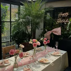 A chic and intimate birthday dinner setup in a glass-enclosed space. The table is styled with a delicate pink runner, matching napkins, and simple glass vases holding pink roses and carnations, with a "Happy Birthday" neon sign in the background.