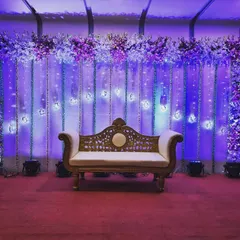 Create a dramatic look with this reception stage rental. It features a classic white sofa set against a backdrop of draped curtains, hanging crystal lights, and vibrant purple uplighting.