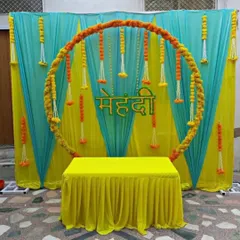 A clean and modern Mehndi setup with a circular marigold ring as the centerpiece. The backdrop uses a combination of turquoise and yellow drapes with hanging floral strings, creating a fresh and bright look.