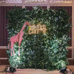 A fun photo booth for the "Wild One" party, featuring a wall of green leaves, a sparkling pink giraffe, and a "Let's Party" neon sign.