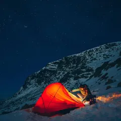 A lone adventurer prepares their glowing tent as night falls on a snowy mountain. The deep blue sky is filled with stars, highlighting the solitude and beauty of winter camping.