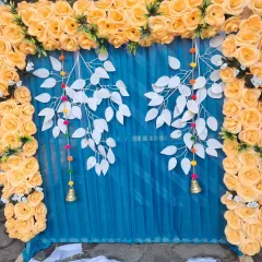 For a festive pooja backdrop, I combined a bright yellow artificial rose frame with a rich blue fabric curtain. The design is accented with decorative white leaf hangings and small bells for a unique and modern look.
