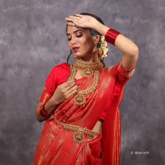 A close-up shot focusing on the emotional aspect of a bridal look. The model's pose draws attention to the intricate necklace and the heavy embroidery of the red saree.