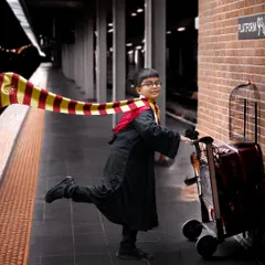 A magical Harry Potter themed photoshoot featuring a young boy in a Gryffindor robe pushing a luggage trolley towards a Platform 9¾ sign on a brick wall.