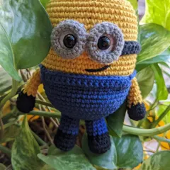 A cheerful Minion toy, crocheted with his classic blue overalls and big grey goggles, hiding among some green leaves.