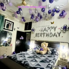A full room view of a first birthday surprise for a wife in her new home. The ceiling is filled with purple and silver balloons, and the bed is decorated with a teddy bear, creating a dreamy, celebratory space.
