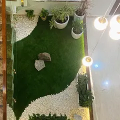 An overhead view of the indoor courtyard garden, showing the careful composition of artificial grass, white pebbles, and natural rocks. The design creates a dynamic, flowing shape that makes the most of the compact space.