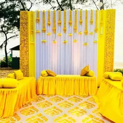 This is an elegant Haldi stage setup with a white and yellow theme. The backdrop features hanging marigold strings against a white curtain, with yellow seating and floor decor to complete the look.