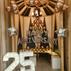 A stylish anniversary party corner. This setup includes a "Happy Anniversary" balloon display, a "25" marquee number, and star-shaped foil balloons inside a wooden gazebo structure.
