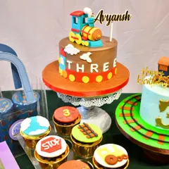 A closer look at the train-themed dessert table, showing the main cake with a fondant train topper, a second smaller cake, and a variety of cupcakes with different train-related designs.