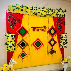 A colorful Haldi backdrop combining red and yellow fabrics for a bold look. The setup is decorated with hanging traditional motifs and bordered with artificial white and yellow flowers, creating a rich and festive frame.