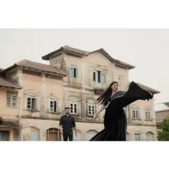 A wide shot showcasing the couple against a grand, old building. The woman's flowing black saree adds a dramatic touch to the scene.