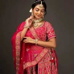 A three-quarter portrait of the bride looking down gracefully, her hands delicately adjusting her necklace. This pose highlights the rich embroidery of her lehenga.