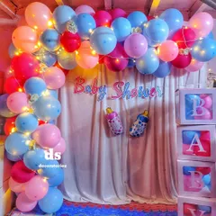 A full view of our pink and blue baby shower balloon decoration. This setup includes a balloon arch woven with fairy lights, a simple curtain backdrop, 'BABY' blocks, and themed foil balloons like the baby bottle.