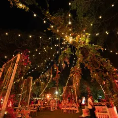An evening view of the Lohri celebration, where strings of fairy lights create a magical canopy over the garden. The warm, rustic setup with wooden tables and chairs is made festive with red-hued floral and vegetable decor.