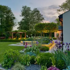 This expansive garden design features a wooden pergola in the background, creating a destination point within the landscape. The structure is subtly lit to draw the eye and provide a sheltered seating area for enjoying the garden at dusk.
