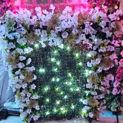 A close-up of the lit green grass wall backdrop. The combination of lush greenery, white and gold flowers, and warm lights creates an elegant and celebratory feel. Book now before it's gone!