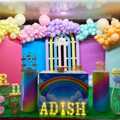 A vibrant rainbow themed birthday party with a colorful block backdrop, a pastel balloon garland, and props like a light-up '1' and a decorative bicycle.