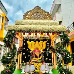 A beautiful Ganesh Chaturthi mantap featuring a large lotus motif made of flowers as the central backdrop. The setup is framed by an arch decorated with white flowers and greenery, creating an elegant space for the Ganesha idol.