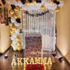 A cozy and romantic birthday surprise at home. This setup features a path of rose petals, marquee letters, and a balloon arch against a fairy-lit curtain.