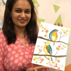 A student beaming with pride as she holds up her delicate watercolor painting of two birds. Learning to use watercolors is a gentle and rewarding process, and her joy is the perfect result.