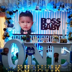 A grand Boss Baby theme for Rudhvik's first birthday celebration. The setup includes a large "ONE" table, multiple character cutouts, and a blue and gold balloon garland against a striped backdrop for a stylish and fun party.