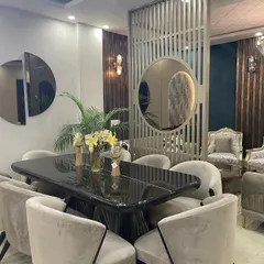 An elegant dining area featuring a large, black marble-top dining table surrounded by plush, beige velvet chairs. A custom slatted wooden partition with a circular mirror adds a contemporary touch and separates the space beautifully.