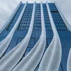 A low-angle view of the calligraphic facade, emphasizing its verticality and the way the perforated panels curve and undulate. This design provides solar shading for the offices below while opening up for residences above.