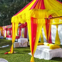 Colorful canopies (shamianas) set up on a lawn for a daytime wedding event. These tents provide seating areas for guests and are decorated in bright yellow and pink fabrics.