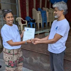 A personal moment of achievement. An instructor presents a new teacher with her certificate and a sacred thread, symbolizing the passing of knowledge and the beginning of her journey as a guide for others.