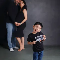 A creative pregnancy announcement photo. The older brother stands in the foreground proudly holding the ultrasound picture, while his parents embrace in the background, creating a fun and memorable family moment.