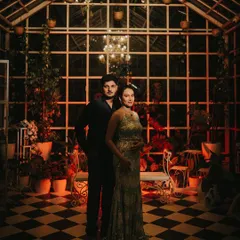 An atmospheric maternity portrait of a couple in a beautifully lit, greenhouse-style room at night. Her shimmering gown and his dark attire create a moody, romantic feel, showing a different, more intimate side of maternity photography.