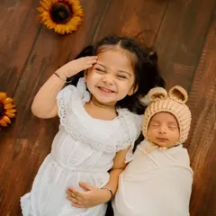 An overhead shot captures a beautiful moment between an older sister and her newborn sibling. Her joyful, protective smile as she lies next to the sleeping baby is simply precious.