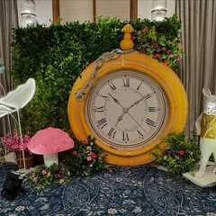 A fantastic photo corner featuring a giant yellow pocket watch prop against a green hedge wall. A white rabbit figure and illuminated butterfly add to the magical, wonderland feel.