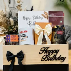 A birthday hamper for him, presented in a rustic wooden crate. It includes a "Vintage 1975" mug, gourmet coffee, and other treats, all styled with a masculine black and cream theme.