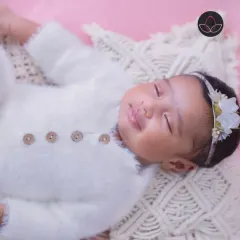 A sleeping newborn with a gentle smile, dressed in a soft white outfit and adorned with a delicate floral headband.