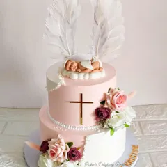 A blessed beginning. This two-tier Baptism cake in pink and white features a sleeping baby with angel wings, a golden cross, and beautiful florals. It's a serene and elegant centerpiece for a sacred day.