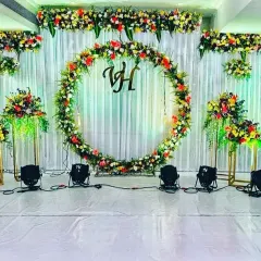 A vibrant and cheerful engagement stage setup. The centerpiece is a large floral ring with the couple's initials, set against a clean white backdrop. The colorful flower arrangements on either side add a festive touch to the celebration.